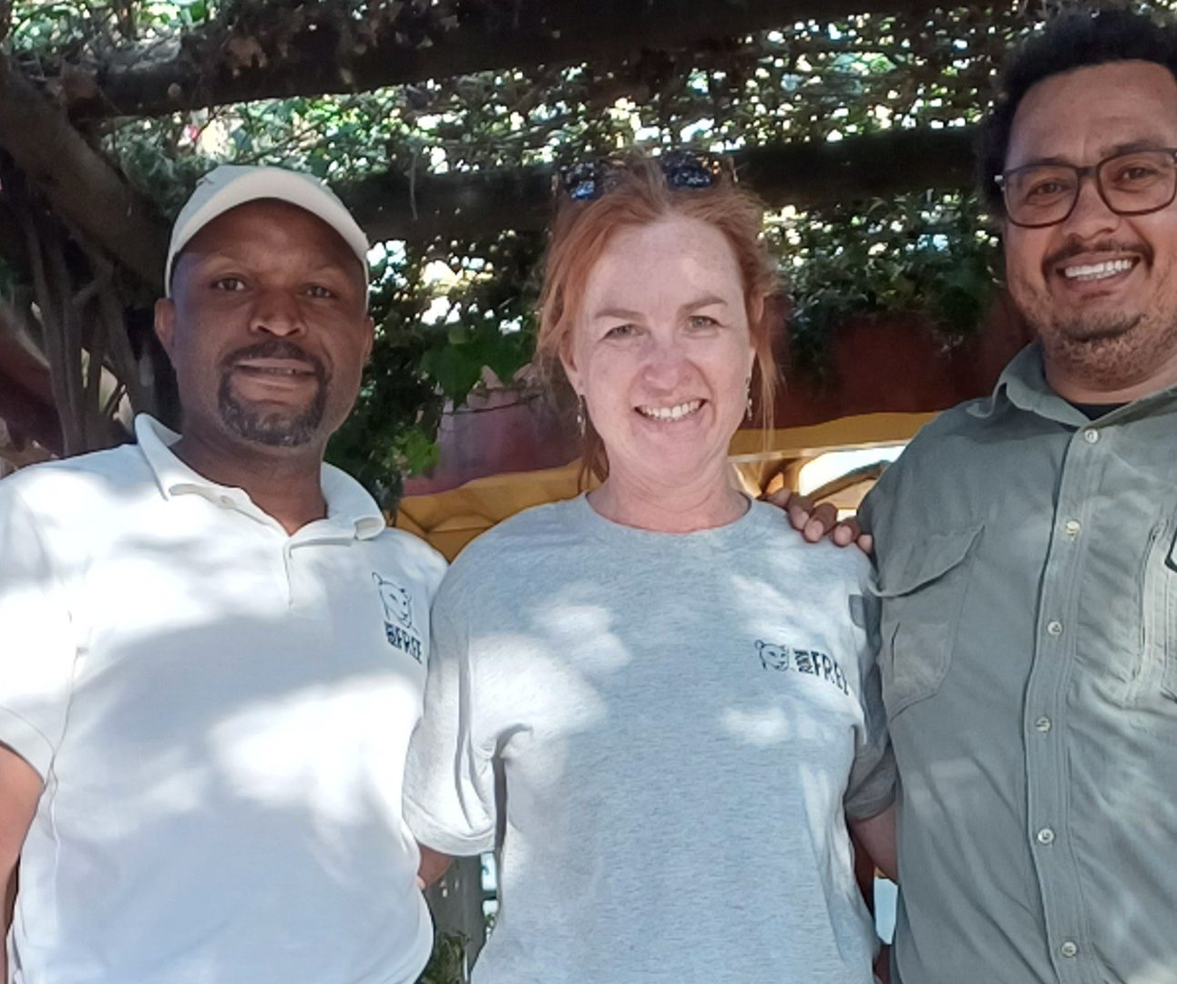 Three people standing with their arms around each other, under a shady pergola