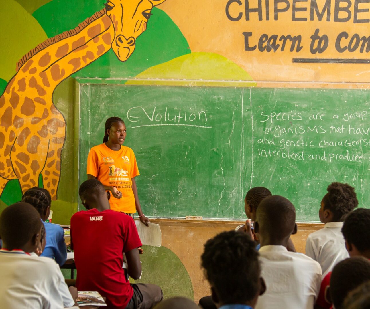 Photograph of a classroom setting with a teacher standing in front of a chalkboard, explaining a lesson on evolution to seated students. The classroom features a large painted giraffe and the phrase 
