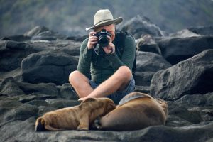 Dave Currey sits cross legged behind two sealions, with a camera raised in front of his face