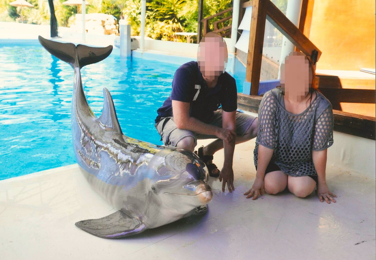 Two people crouching down next to the side of a pool, next to a dolphin who is being made to pose for a photo.