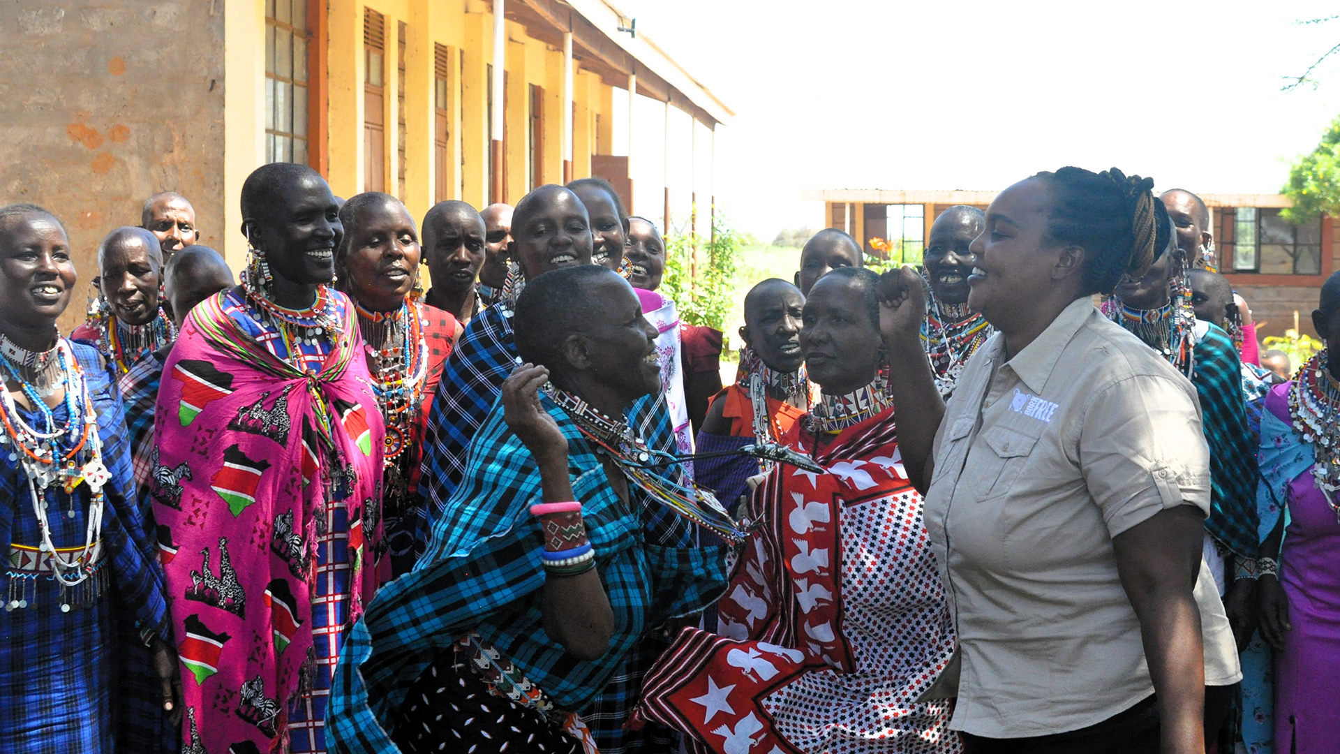 A Kenyan woman in traditional dress is leaning forward and dancing with a woman in Born Free uniform, in front of a crowd