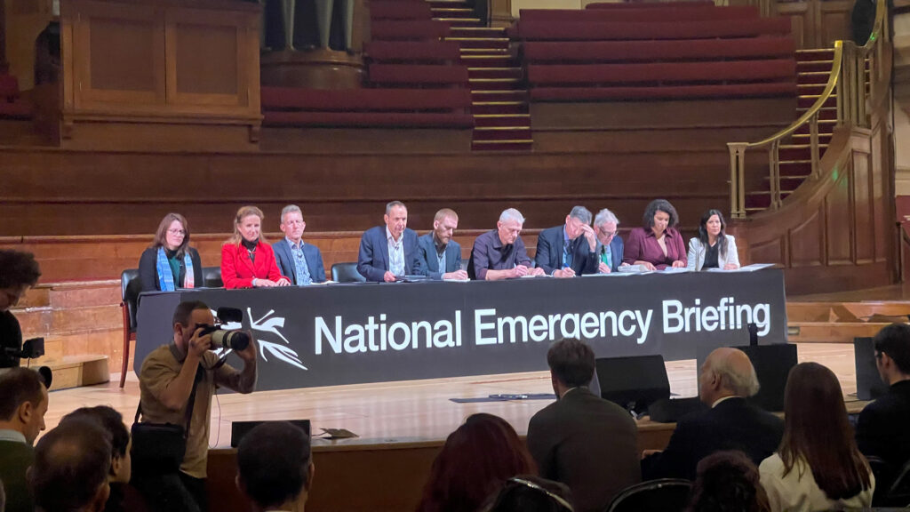 A panel of people sit behind a long desk on a stage, a banner reads National Emergency Briefing