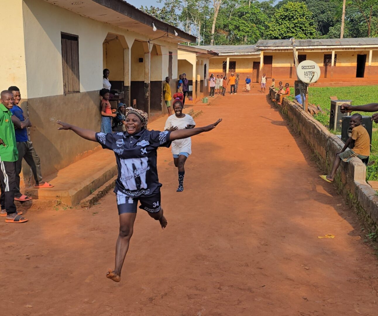 Photograph showing a group of women taking part in a running race, and one woman joyfully crossing the finish line. The scene features vibrant clothing, joyful expressions, and a mix of standing and moving figures, highlighting a lively outdoor activity.