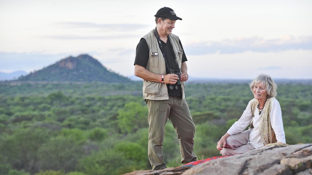 Will Travers stood next to Virginia McKenna, who is sitting down on a rock with a mountainous Kenyan landscape in the background
